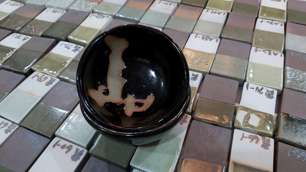 路慶麟用電瓦斯窯燒製古青瓷釉藥試片，以及在台藝大時期的茶碗作品，碗內有他的書法行草字體「天地」。（圖/陳惠玲攝影）